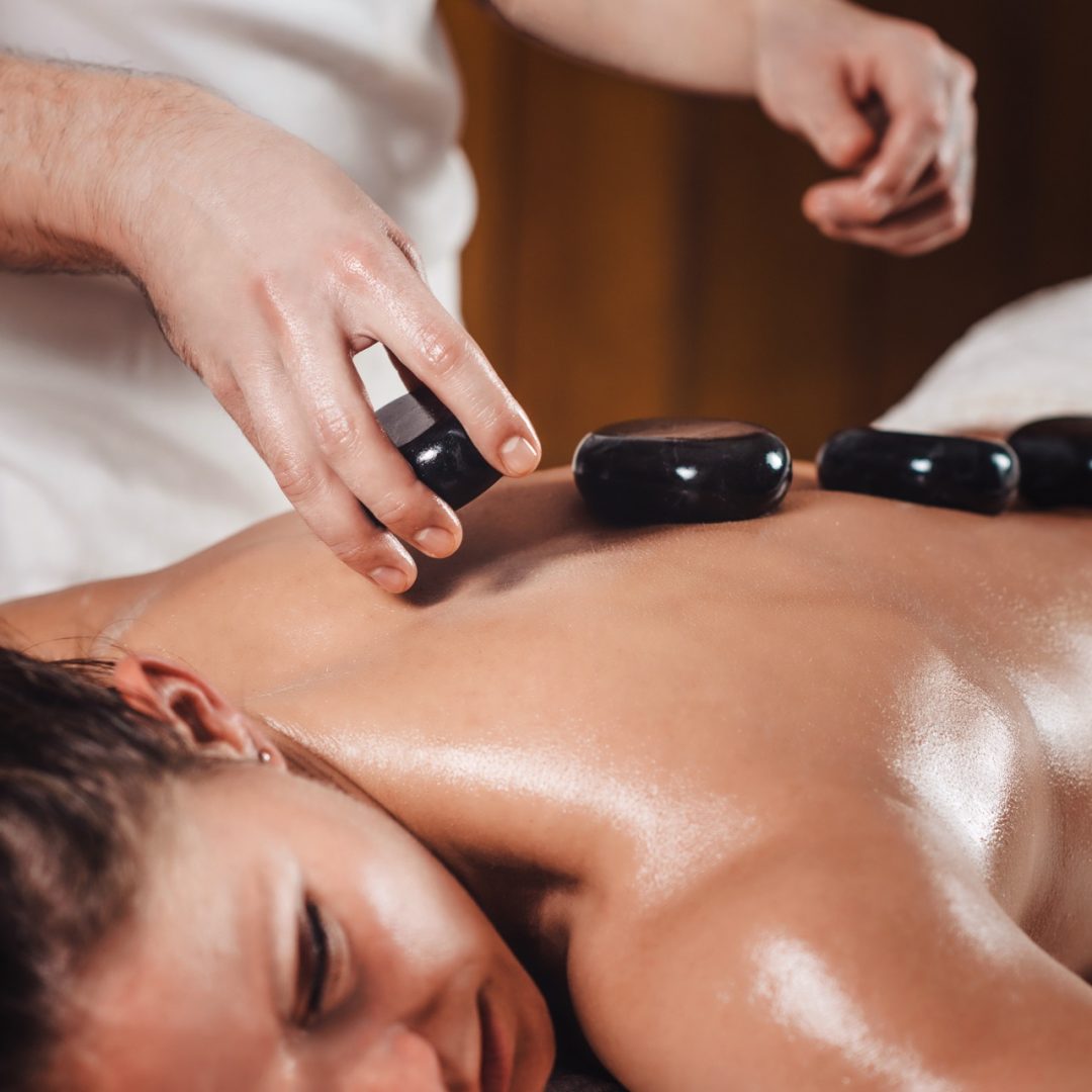 Beautiful young woman getting hot stone massage at spa salon, lying on massage table